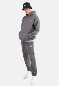 Grau Hoodie mit Fronttasche und Logo, passende graue Jogginghose mit Kordelzug, kombiniert mit weißen Sneakers und einem schwarzen Bucket Hat.