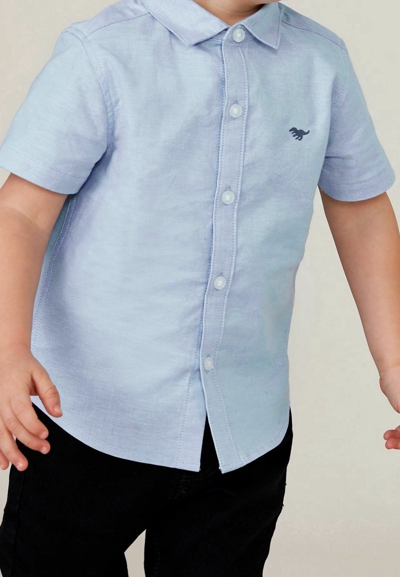 Tout-petit portant une chemise à manches courtes bleu clair avec un petit logo dinosaure et un pantalon noir, bras légèrement tendus.