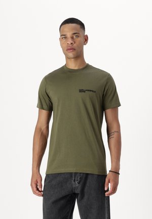 Camiseta de algodón verde oliva de mangas cortas, con cuello redondo y un logo negro impreso en el lado izquierdo del pecho. Combinada con jeans gris oscuro.