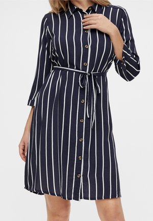 Vrouw draagt een marineblauwe jurk met witte verticale strepen, knoopsluiting aan de voorkant, tailleband en driekwart mouwen.