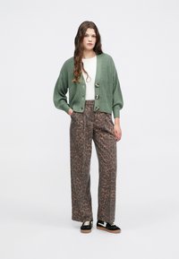 Zaļa adīta jaka ar trim lielām pogām, apvienota ar platām brūnām biksēm ar melnu leoparda rakstu. Melni sporta apavi ar brūnu zoli.