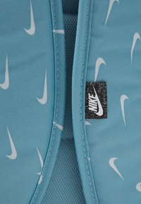 Blå Nike-ryggsäck med en slät tygtextur som har upprepade vita swoosh-loggor och en svart Nike-märke prydd med vit text.