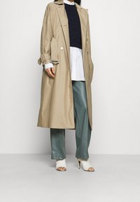 Trench coat beige con cintura, maniche lunghe e chiusura a bottoni, indossato sopra un maglione blu navy e una camicia bianca, abbinato a pantaloni verdi e tacchi bianchi.