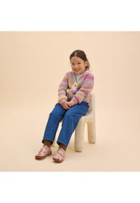 Multikleurige gebreide cardigan met roze, gele en lavendelkleurige strepen, denim jeans en roze bloemen instappers, op een textuur witte stoel.