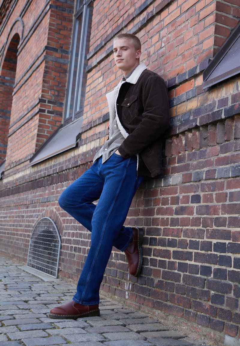 Chaqueta de borrego marrón de corduroy con cuello de pelo sintético, camisa gris, jeans azules y botas de cuero burdeos, posando frente a una pared de ladrillo.