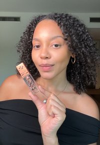 Produit de maquillage liquide clair dans un tube transparent avec une étiquette dorée et noire, tenu par une personne aux cheveux bouclés, portant un haut noir.