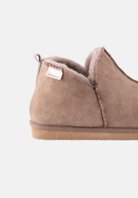 Pantofole in suede marrone con un interno morbido e dettagli cuciti. Presentano una forma arrotondata e una suola in gomma per una buona aderenza. Etichetta visibile sul lato.