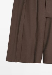 Pantalon marron foncé sur mesure, coupe large, plis marqués et détail passant de ceinture à la taille.
