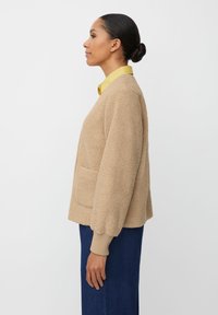 Beżowa, teksturowana kurtka z polaru z długimi rękawami, ściągaczami na mankietach i przednią kieszenią. Noszona na żółtej koszuli z kołnierzykiem, o luźnym kroju.