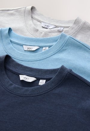 Drie katoenen t-shirts gestapeld, in lichtgrijs, zachtblauw en marineblauw. Elk shirt heeft een ronde hals en een label met het merk "NEXT."