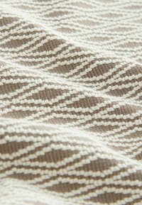 Textiel met een golvend patroon in beige en crème, met een gestructureerd breiwerkontwerp. Het oppervlak toont afwisselend gladde en verhoogde steken in rijen.