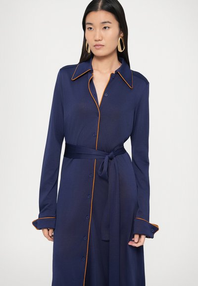 Φόρεμα σε ναυτικό μπλε με πορτοκαλί λεπτομέρειες, κουμπιά, μακριά μανίκια και δεμένη ζώνη στη μέση. Λείο ύφασμα με άνετη εφαρμογή και γιακά.