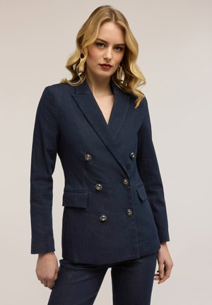 Kvinde med blondt hår iført en marineblå dobbeltradet blazer med gyldne knapper og matchende bukser mod en ensfarvet lys baggrund.