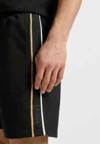 Zwarte sportshorts met een gestructureerde tailleband, zijstrepen in wit en goud, en een subtiele logo-afdruk op de zoom.