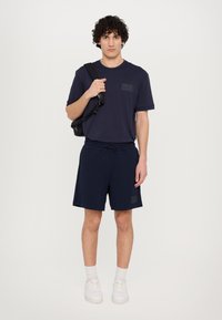 Marineblå atletisk t-shirt med en brystlomme, parret med marineblå shorts med elastisk talje og snor. Hvide sneakers iført.