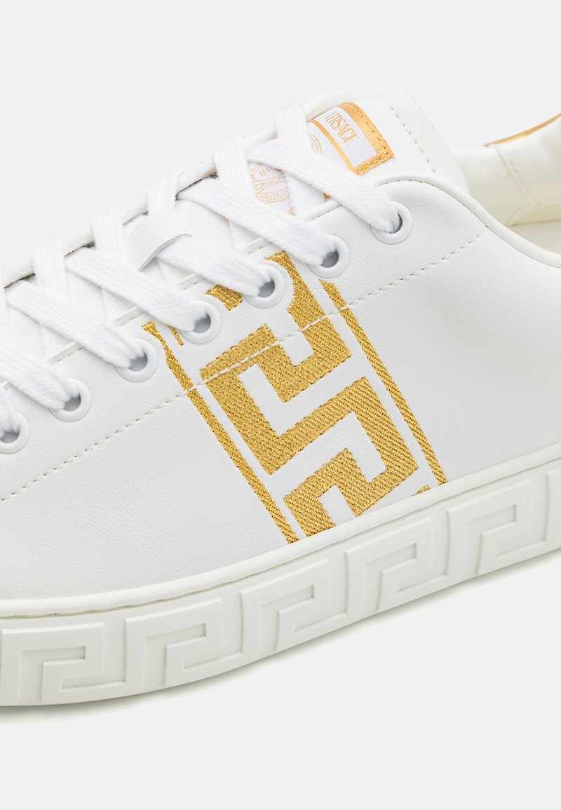 Sneaker bianchi in pelle con un motivo geometrico dorato testurizzato, lacci bianchi e suola in gomma con motivo. Etichetta con il marchio dorato ben visibile.