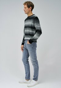 Suéter de punto a rayas gris y negro con cuello redondo, combinado con jeans gris claro y zapatillas blancas. La modelo posa de perfil.