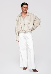 Mujer con cabello oscuro y gafas, vistiendo una chaqueta beige con cremallera y cinturón, pantalones blancos de pierna ancha y tacones puntiagudos marrones, posando con la mano en la cadera.