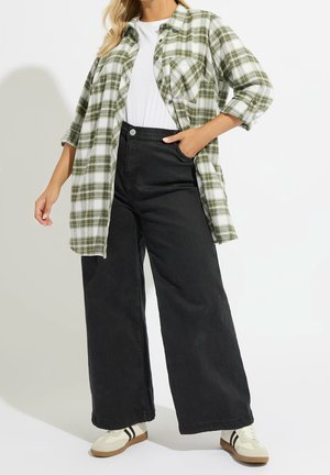 Kvinde iført hvid skjorte, grøn ternet åben knap-på, viddebukser i sort og beige sneakers med sorte striber, stående mod en hvid baggrund.