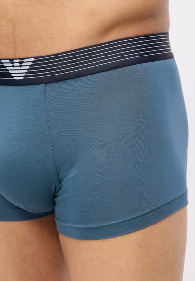 Κοντινό πλάνο ενός ατόμου που φοράει μπλε μποξεράκια με μαύρη και λευκή ριγέ μέση που διαθέτει λογότυπο.