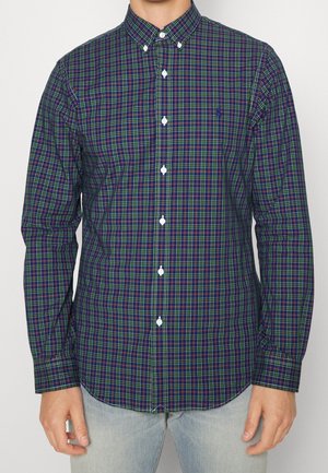 Mand iført en langærmet skjorte med navy, grøn og rød tern med knapper og lyseblå jeans, stående mod en ensfarvet baggrund.