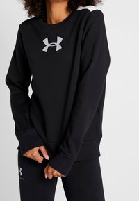 Sudadera negra con cuello redondo, puños de canalé y un gran logo gris en el pecho. La tela parece suave y ligeramente texturizada.