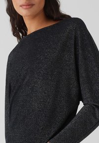 Schwarzes Oberteil mit weitem Ausschnitt, glitzernder Textur und langen Ärmeln. Der Stoff hat einen dezenten Schimmer für zusätzliche visuelle Akzente.