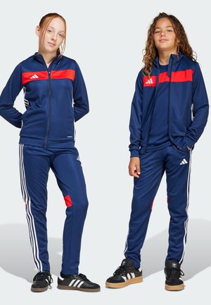 Marine tracksuits met rode accenten, gemaakt van gladde stof. Witte gestreepte details langs de broek. Zwarte sneakers met rubberen zolen.