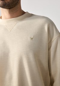 Beige langermet skjorte laget av teksturert stoff, med rund hals og et lite gullfuglemerke på venstre brystområde.