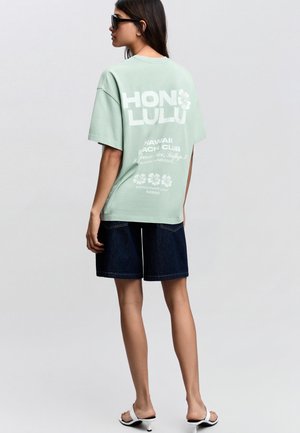 Kobieta w miętowo-zielonej, oversize'owej koszulce z napisem "HONOLULU" i kwiatowymi nadrukami, ciemnych szortach jeansowych, białych sandałach i czarnych okularach przeciwsłonecznych.
