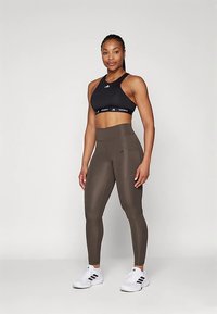 Sujetador deportivo negro con logo, leggings verde oliva con bolsillos y zapatillas deportivas blancas con detalles negros. Diseño y textura lisos y ajustados.