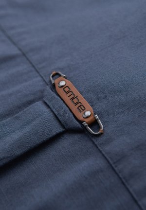 Étiquette de vêtement en cuir marron avec texte noir "ombre" attachée à un tissu texturé bleu foncé avec des rivets et crochets en métal.