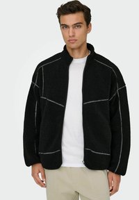 Veste en polaire noire avec fermeture éclair sur le devant, présentant des coutures grises contrastantes, un col haut, des manches larges et des poches latérales.