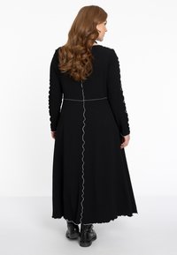 Schwarzes Langarmkleid aus strukturiertem Stoff, mit Rüschensaum und kontrastierenden weißen Nähten entlang der Nähte. Getragen mit schwarzen Stiefeln.