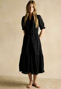 Schwarzes, kurzärmliges, schichtweises Midi-Kleid mit Knopfverschluss und Kordelzug in der Taille. Augenmuster und lockere Passform. Getragen mit leichten Sandalen.