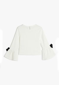 Top bianco con texture, caratterizzato da maniche lunghe a campana con dettagli in nastro nero ai polsini e una piccola apertura sul retro. Design semplice e cropped.