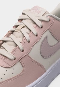 Κοντινό πλάνο από μπεζ και ροζ παπούτσι Nike Air Force 1 με λευκά κορδόνια και υφασμάτινη δερμάτινη λεπτομέρεια στο πλάι.