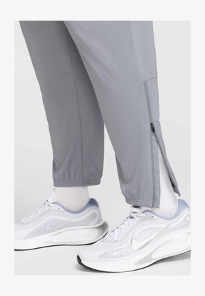 Pantaloni sportivi grigi con cerniere alle caviglie, abbinati a scarpe da corsa bianche e argentate e calze bianche. Tessuto testurizzato, vestibilità affusolata.