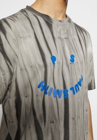 Camiseta de algodón en tie-dye gris, con un gráfico de una cara sonriente azul y el texto "PAUL SMITH" en letras en negrita. Mangas cortas y cuello redondo.