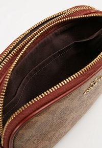 Sac en tissu à motif marron avec une fermeture éclair bordeaux et une doublure assortie. L'intérieur présente un textile marron lisse. Accents de fermeture éclair dorés.