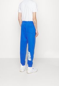 Kungablå joggingbyxor med elastiskt midjeband och sidoficka, med "JORDAN" tryckt i vitt längs vänster ben. Gummiaktig textur.