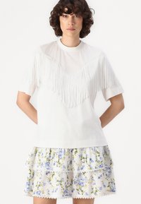 Weißes T-Shirt mit Fransenverzierung auf der Vorderseite und kurzen Ärmeln, kombiniert mit einem fließenden, mehrlagigen Rock mit Blumenmuster in Blau- und Grüntönen.