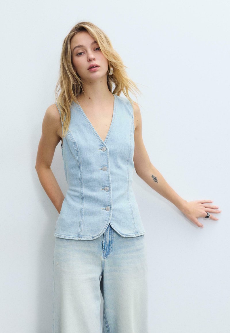 Mladá žena s dlhými blond vlasmi má na sebe svetlomodrú bezrukávovú vestu na gombíky a k nim ladia široké džínsové nohavice, stojí opretá o bielu stenu.