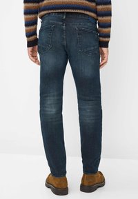Uomo che indossa jeans blu scuro, stivali di camoscio marroni e un maglione a righe, in piedi con le mani rilassate ai fianchi, rivolto di spalle.