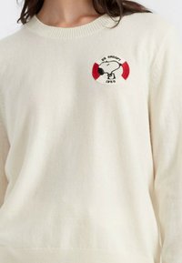 Crème-kleurige trui van zachte stof, met een ronde geborduurde patch met een cartoonfiguur en kleurrijke accenten.