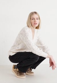 Maglione lavorato a maglia crema con motivo floreale, scollatura a V e polsini a costine, abbinato a pantaloni scuri e sneakers beige con dettagli in pelliccia.