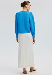 Blusa blu con maniche a sbuffo e bottoni decorativi, abbinata a una gonna maxi bianca e texturizzata. L'outfit presenta una silhouette pulita e moderna.