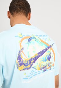 Světle modré tričko s barevným grafickým motivem na zadní straně, zobrazením mořských želv, bublinek a abstraktního designu s živými barvami.