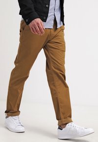 Braune Baumwoll-Chinos mit schmaler Passform, ausgestattet mit Seitentaschen, hochgekrempelten Säumen, kombiniert mit weißen Turnschuhen. Glatte Textur und sauberes Design.