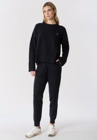 Maglione nero lavorato a maglia con orlo a coste, abbinato a pantaloni jogger neri coordinati. Il modello indossa scarpe da ginnastica bianche. Design semplice e casual.